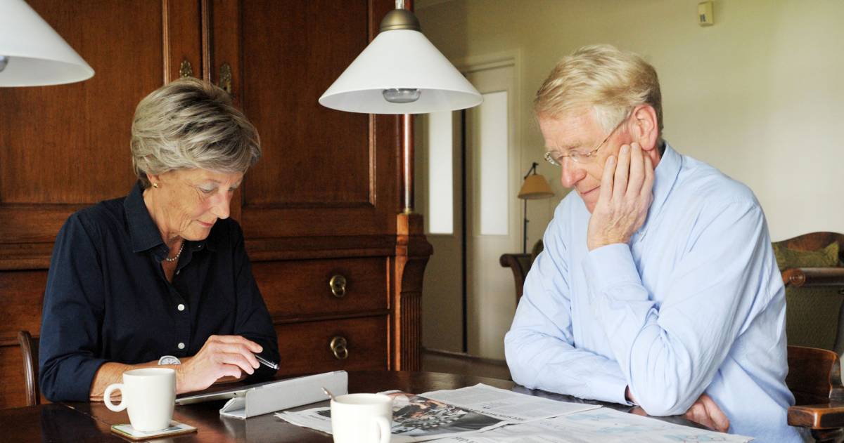 Een ouder stel dat aan tafel zit, de man leest de krant, de vrouw bekijkt iets op haar tablet