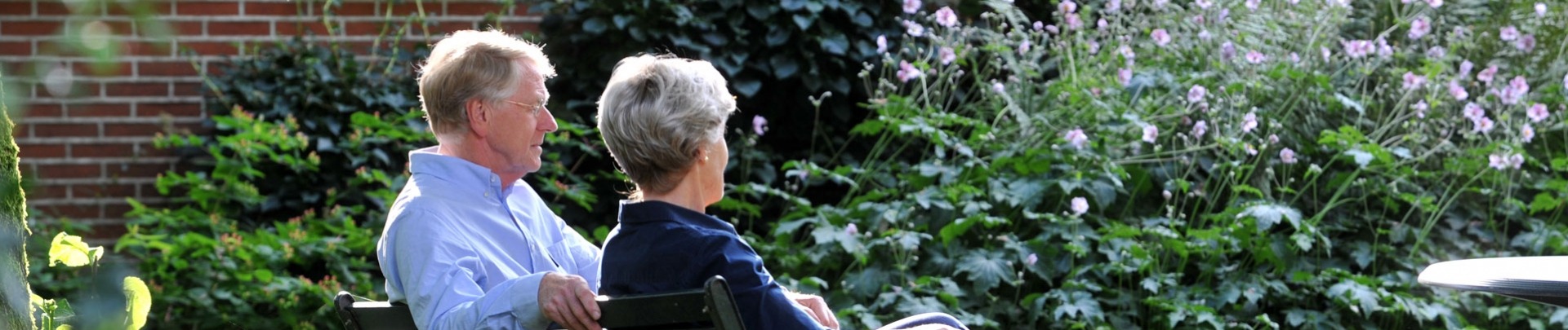 Een ouder stel zit buiten in de tuin op een bankje en ze kijken van zich af