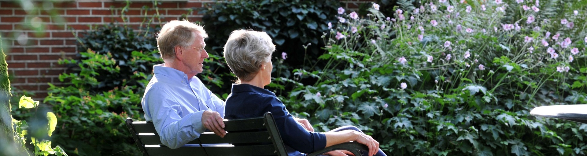 Een ouder stel zit buiten in de tuin op een bankje en ze kijken van zich af